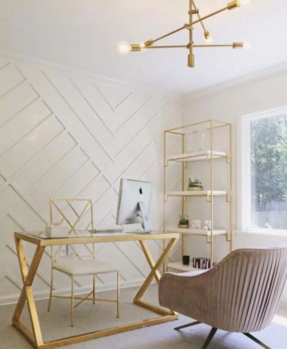 a glam home office with a white paneled geometric wall, a gold etagere, desk and chair, a blush chair and a bold chandelier