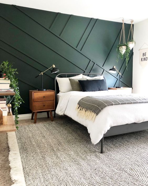 a mid-centiry modern bedroom with a dark green geometric panel wall, a metal bed, wooden furniture and some greenery in pots to enliven it